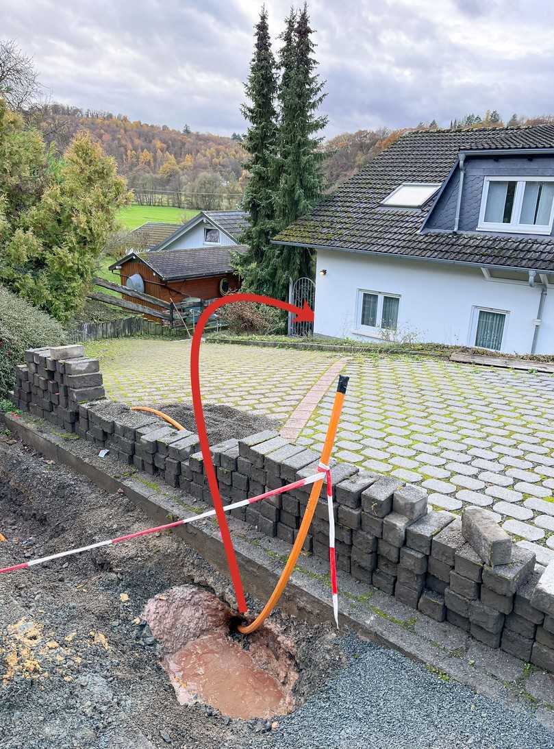 Blick auf das eingezogene Verbundrohr und die abschüssige Einfahrt des Grundstückes mit der Hausecke, um die herum in einem Winkel von 80° gebohrt werden musste. Der rote Pfeil markiert die Bohrrichtung. | Foto: L-Team X-Tools